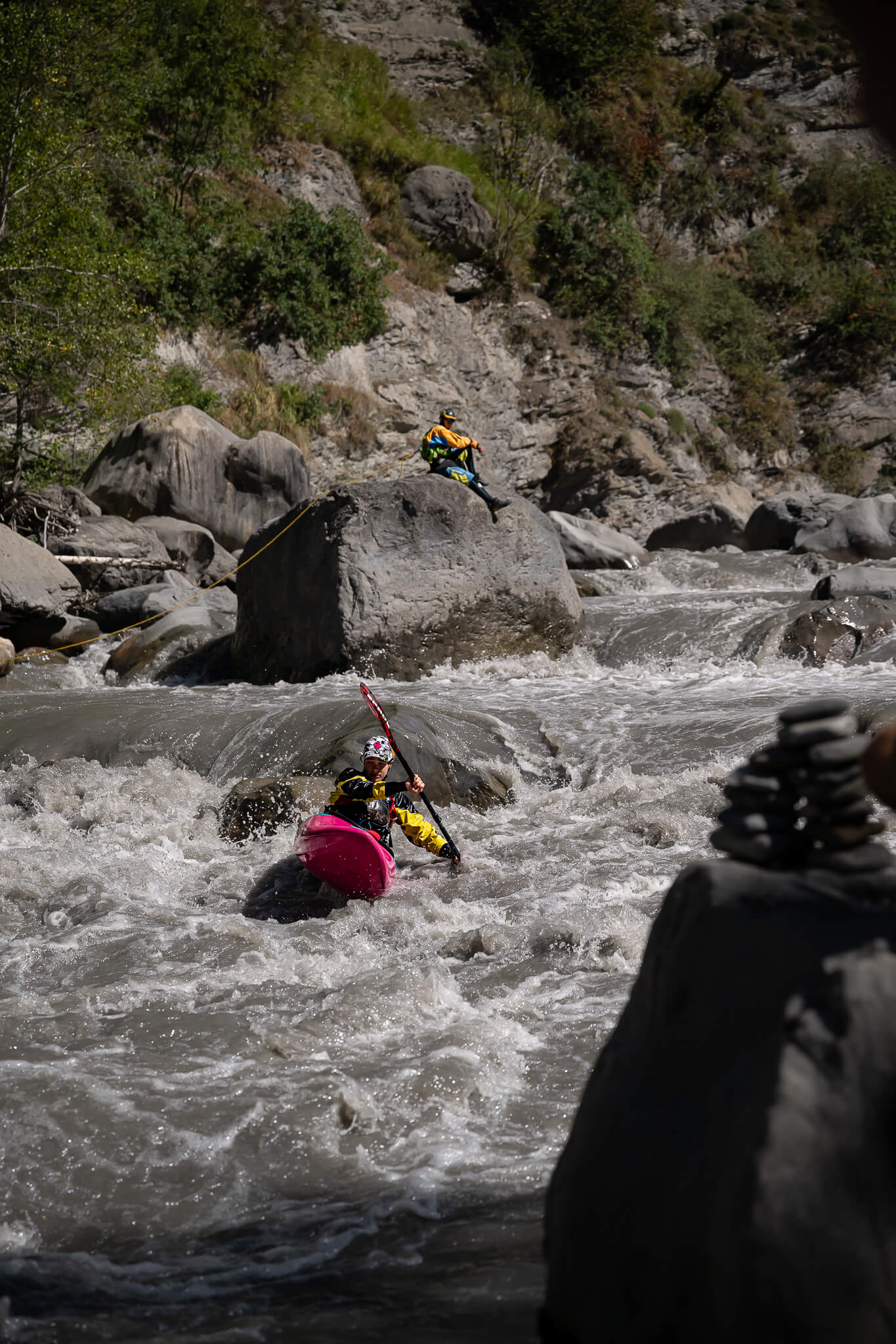 ubayak-03-same-position-200mm-cedric-eyma A racer dropping through the rapid beside a large boulder, a stone cairn in the foreground, shot at 200 mm.